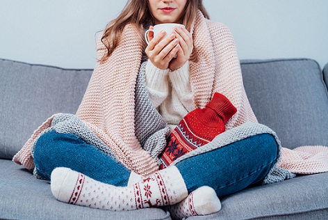 אישה קופאת בחורף. ילדה צעירה לובשת גרבי צמר חמות ועטופה בשתי שמיכות, מחזיקה כוס שתייה חמה וכרית חימום כשהיא יושבת על הספה בבית. לשמור על חום. מיקוד סלקטיבי.